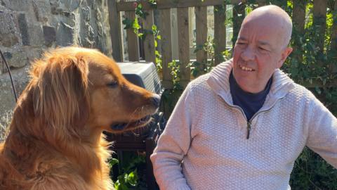 A man wearing a light-coloured jumper is looking at a dog who is sat to the side of him. They are outdoors, in what appears to be a garden with a fence nearby behind them.