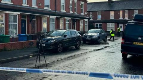 Police tape across a residential street in Moss Side. There is a camera on a tripod in the road, and two police officers in uniform in the backdrop. A bicycle is on its side next to a car within the cordon. 