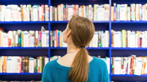 Rear view of a person with long brown hair in a ponytail, looking at bookshelves filled with books on vibrant blue shelves.