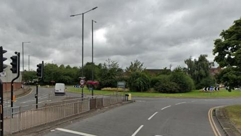 A roundabout, featuring a large island with trees. Traffic lights, a pedestrian crossing and other street furniture can also be seen.