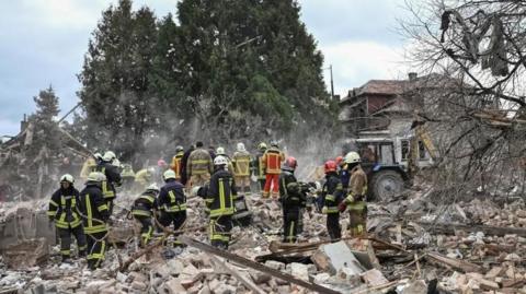 A number of rescuers in protective clothing can be seen working around a destroyed house. They are surrounded by rubble and are clearing away bricks and other pieces of debris.