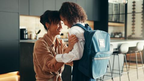 A parent kneels in a modern kitchen, holding and comforting a school‑aged child who is wearing a uniform and a large backpack, as they share a close and supportive moment.