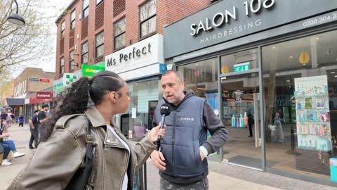 A woman wearing a leather jacket interviews a man in a padded vest outside a hairdressing shop in a busy urban street.