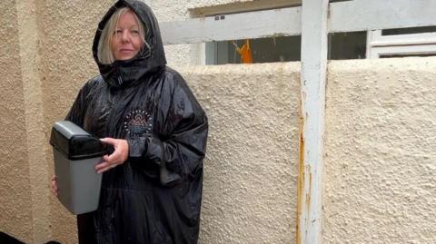 Becci Hey is pictured. She is wearing a black raincoat with the hood up and she has blonde hair. She is holding a small sanitary bin and is standing outside a cream-coloured toilet block.