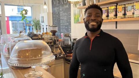 A man with dark hair and beard, wearing a black top, stands behind a coffee shop counter. 