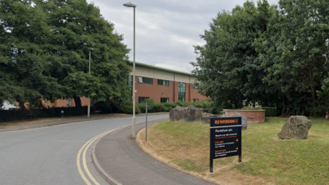 A curved road with double yellow lines leads towards a partially obscured building surrounded by trees. On the right, a grassy verge features large rocks and a black sign reading “RENSHAW” in orange, with “Renishaw plc” and contact details in white beneath.