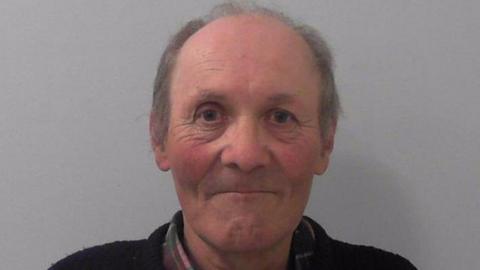 Phipps' mugshot, where he is smiling with thin pursed lips and rosy cheeks. He is balding and has grey hair. He is wearing a tartan shirt with a dark jumper or cardigan over it and is stood in front of a grey background. 