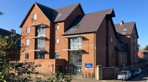 A four-storey redbrick building with a sloped brown roof and large windows.