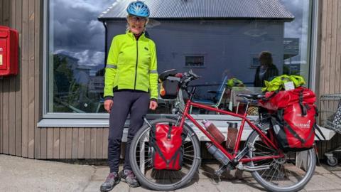A smiling female wearing glasses, a blue cycle helmet, a yellow high visibility jacket and black trousers. She is standing in front of a shop which has a reflection of a building in its large window. The window is surrounded by wooden cladding. 
She is standing next to her bike which has a red frame, a number of water bottles attached to it, and red and black bags loaded on to it. 
