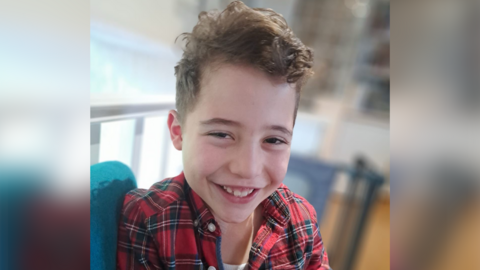 Smiling young boy. He is wearing a chequered shirt and is looking at the camera. He has brown hair.