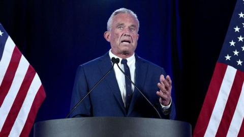 RFK Jr behind a podium flanked by American flags