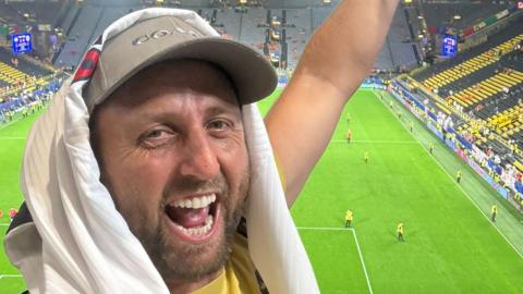 Alex Fox looking into the camera smiling in front of the pitch wearing a cap