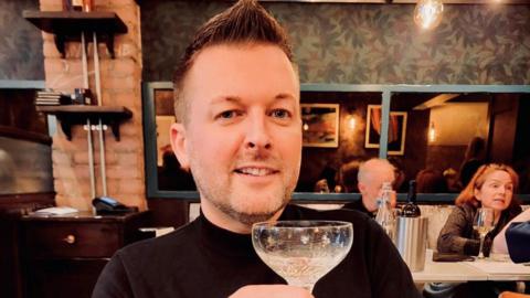 A head and shoulders photo of a Jason Johnson. He has gelled brown hair and short stubble. He is wearing a black jumper and holding an ornate cocktail glass. People can be seen drinking at tables in the background.