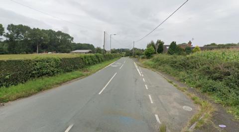 Offoxey Road. A grey road with white markings down the centre and on the right side. On either side of the road are green hedges, fields and trees. 