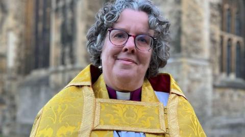 Reverend Canon Mary Gregory smiling at the camera wearing black framed glasses and a yellow robe.