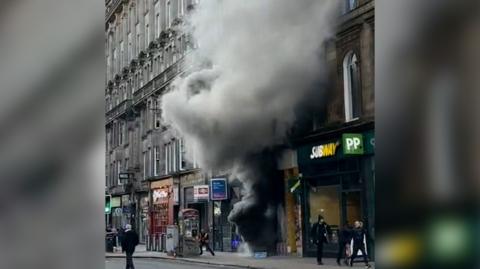 Still showing plumes of grey smoke emitting from a retail shop on Union Street with bystanders looking on