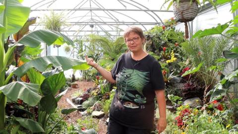 A woman with brown hair and glasses wearing a black T-shirt with different types of green leaves on it. She is standing in a giant green house full of different plants and shrubs.