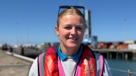 Woman in her 20's wearing sunglasses on her head wearing a pink high vis and a lie jacket. She is stood with St Peter Port Harbour in the background. 
