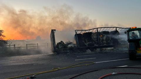 A burned-out lorry lies on its side across a road at dusk, still smoking as water is sprayed on to the smouldering load.