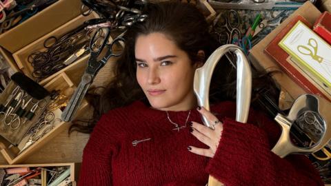 A woman who is a dark red jumper is lying on the floor and is surrounded by scissors. she is looking directly into the camera and is holding a giant pair of silver scissors.