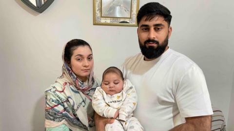 Kamran Agha holds Azlaan in his arms, with the baby's mother Javeria Arooj stood next to them. Javeria is wearing a headscarf and wears a neutral expression. Kamran, who has a black beard and black hair, also has a neutral expression. Azlaan, who has a tube protruding from his nose, is asleep.