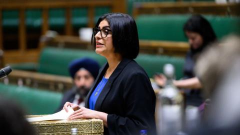 Home Secretary Shabana Mahmood in the Commons. She has shoulder-length black hair and is wearing a black suit jacket and blue shirt. A few other MPs are visible in the blurred background.