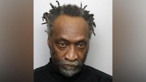 A mugshot of a man staring blankly and wearing a black roll-neck jumper. He has a beard and grey hair in dreadlocks. 