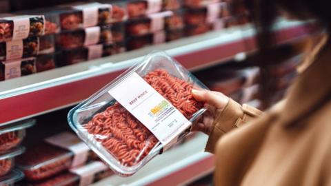 A woman holds a packet of beef mince in a supermarket with rows of mince on shelves in the background