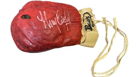 A damaged red boxing glove worn by Muhammad Ali with a white signature is seen against a white background.