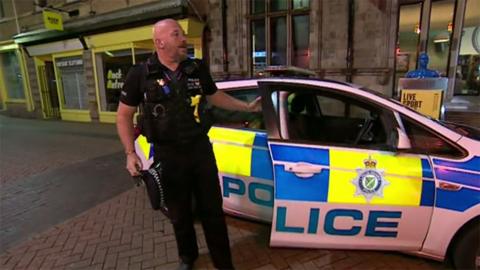 Inspector Jonathan Mellor in a police uniform getting out of a marked police car in Lincolnshire street. He has a shaved head and dark beard he is carrying his police cap in his right hand