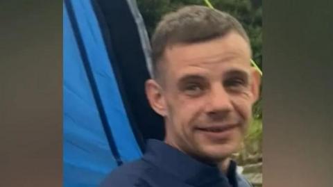 Rikki Berry, 36, who has light brown hair and wears a blue shirt, smiles at the camera with a blue tent and a grassy area in the back-drop. 