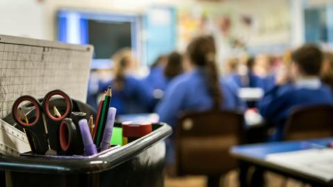 Classroom scene with stationery box in the foreground and blurred images of children with blue school uniform sweaters in the background.