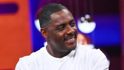 Sir Idris Elba looks away from the camera in a white shirt and smiles. The background is blurred.