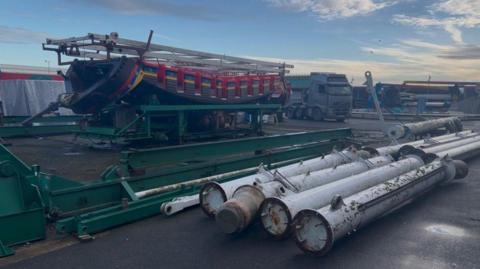 Large metal rods and pieces of a former pirate ship are dismantled and lie on the ground at an outside fairground site