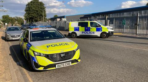 The road at an industrial estate with a police car and a police van parked up. There are metal railings and warehouses behind them, and trees dotted in the distance.
