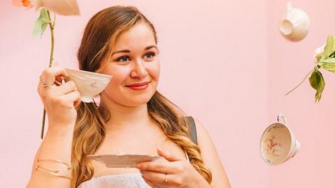 A woman holds a cup in front of a pink background