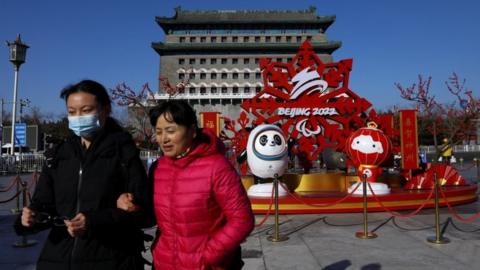 Two women bundled up in winter coats walk past a temple and Olympics display in Beijing