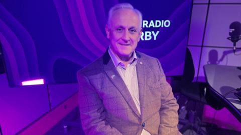 Man in brown tweed jacket with short grey hair sat in radio studio infront of purple background with BBC Radio Derby logo on 