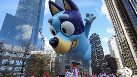 A giant balloon of Bluey hangs from strings in New York city, being pulled along my people wearing red vests and hats.
