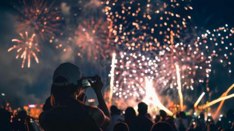 A stock image the back of a girl in a baseball cap filming a fireworks display on her mobile phone.