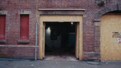 The outside of a red brick building. Windows are boarded up. An entrance is open revealing a bare dusty room inside