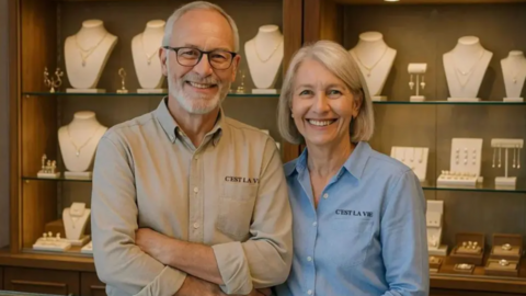 A life-like middle-aged man and a woman are smiling and standing in front of a counter filled with jewellery. They are wearing shirts with the C'est La Vie logo on