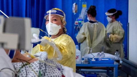 A nurse in PPE looks at a monitor in an ICU with two nurses in the background