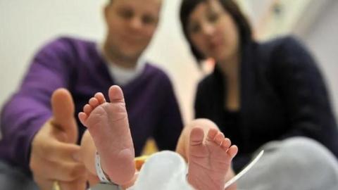 Closeup of a baby's feet with a hospital ID bracelet on the ankle on the left, with a man and a woman out of focus in the background looking down at them. He is wearing a purple jumper and has a gold ring on his left hand and she has dark hair and is wearing a black blazer.