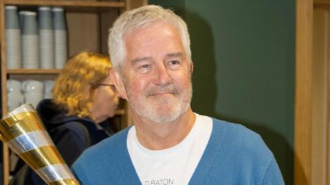 A man with grey hair and a grey beard holding a silver and gold baton while smiling. He is wearing a blue cardigan over a white round-kneck T-shirt.