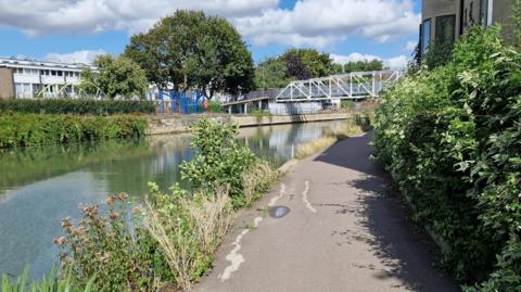 Sir Geoffrey Arthur Building is on the right, with a grey tarmac towpath and greenery lining the River Thames and another building on the left hand side, there is a bridge with white piping going across the river