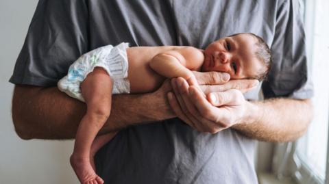 A man with a grey T-shirt holding a baby in his arms. The baby is laying on its belly with its head on the man's palm.