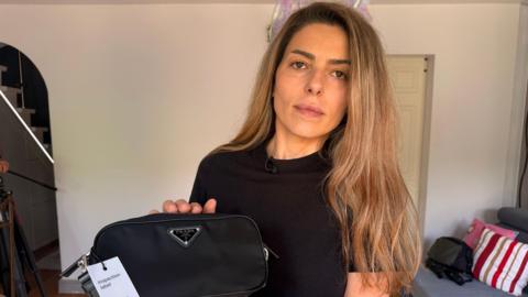 A woman with long dark blond hair holding a black handbag with the Prada logo on it which is fake. She is standing in a living room in a house. It has a grey sofa and multi-coloured cushions. An open staircase can be seen in the background too. She looks serious.