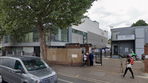 Street view image of a mosque. It is a grey building set back from a road, with tall trees on the pavement outside. There are people walking past and gathered near the mosque.