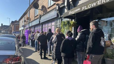 A queue of people on a pavement outside a convenience store. About 13 people can be seen queuing, with an arch of purple balloons outside the shop.